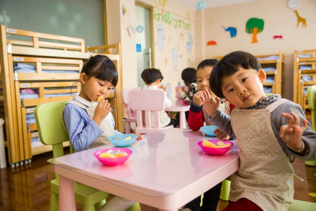 pexels photo 1001914 1001914 1 Kids seated around a table in a colorful classroom, eating snacks happily.